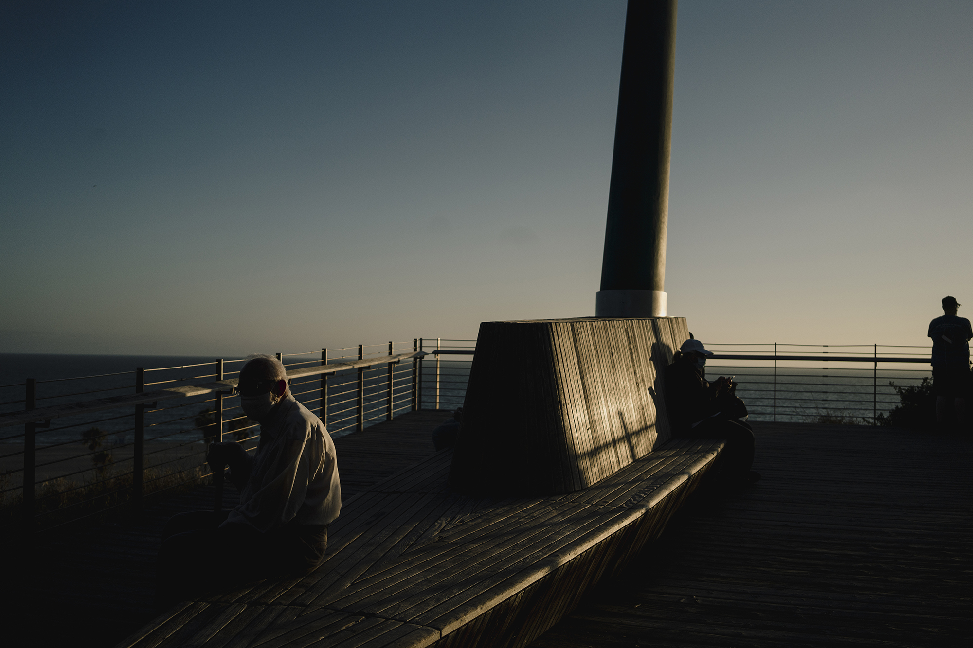 Several people sit or stand on a wooden deck near a railing and a tall column, silhouetted against the sky and ocean.