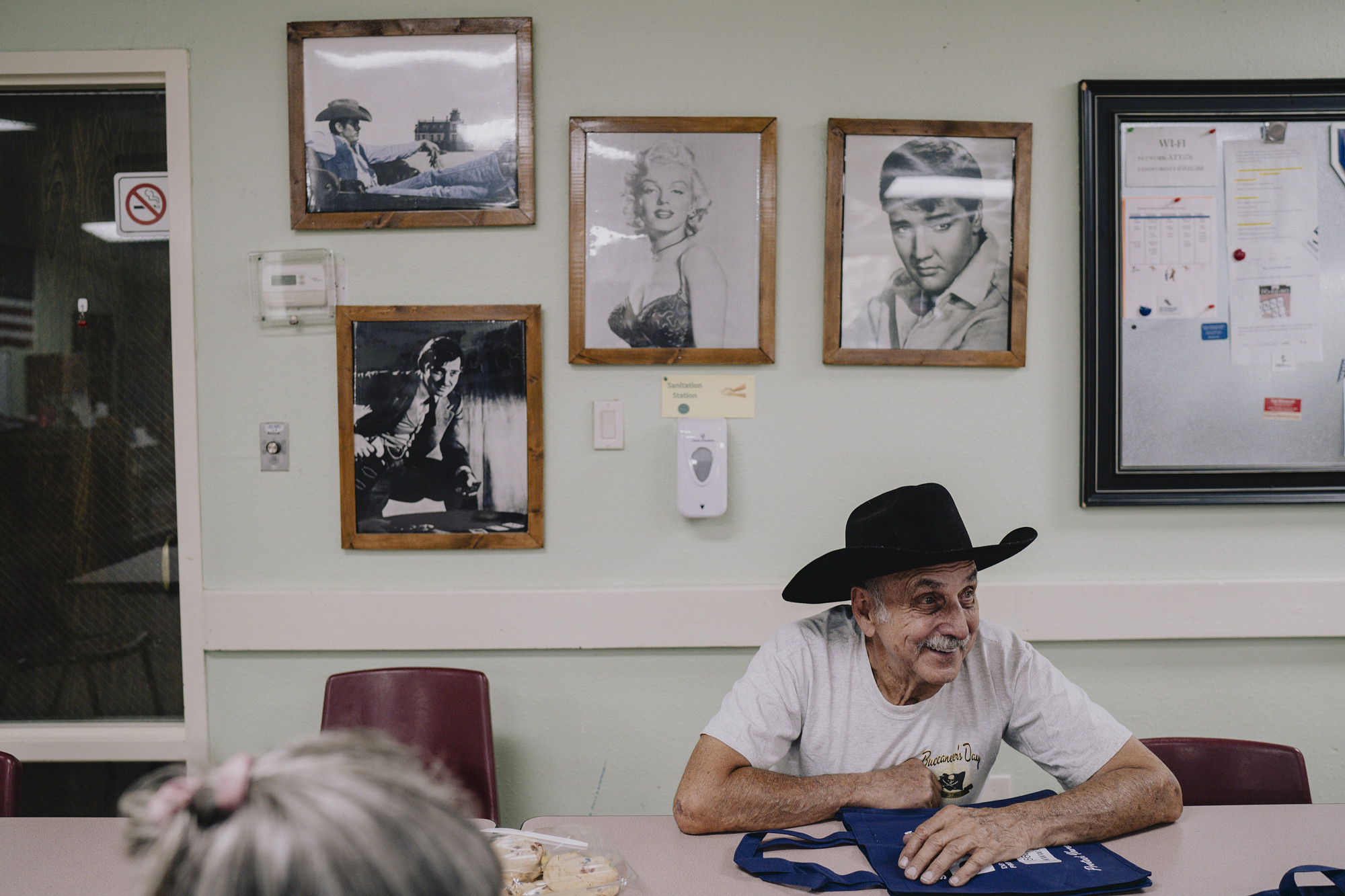 A person wearing a black cowboy hat sits at a table indoors, smiling, with framed black-and-white portraits of celebrities on the wall behind them and a bulletin board to the side.