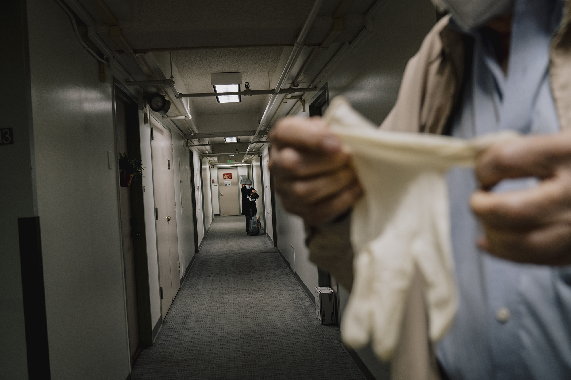 A hallway lined with doors and overhead pipes extends into the distance as a person in the foreground stretches a pair of gloves while another person stands farther down the corridor near a cart.