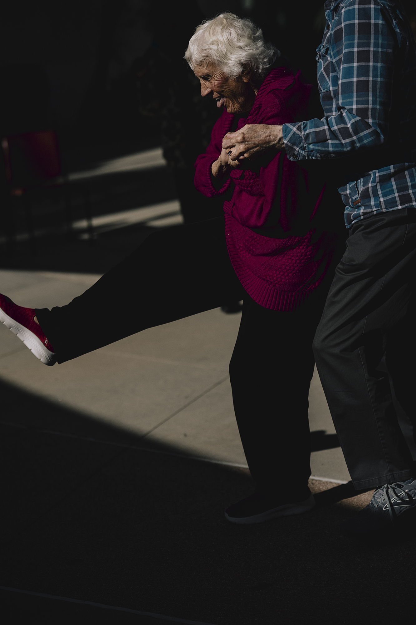 An elderly person in a red sweater lifts one leg while holding onto another person’s arm for support, standing in a patch of sunlight against a darker background.