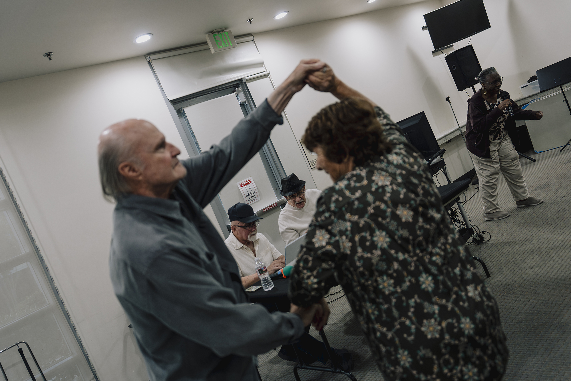 Two people dance together indoors with hands raised while others sit at a table behind them and another person stands nearby holding a microphone.
