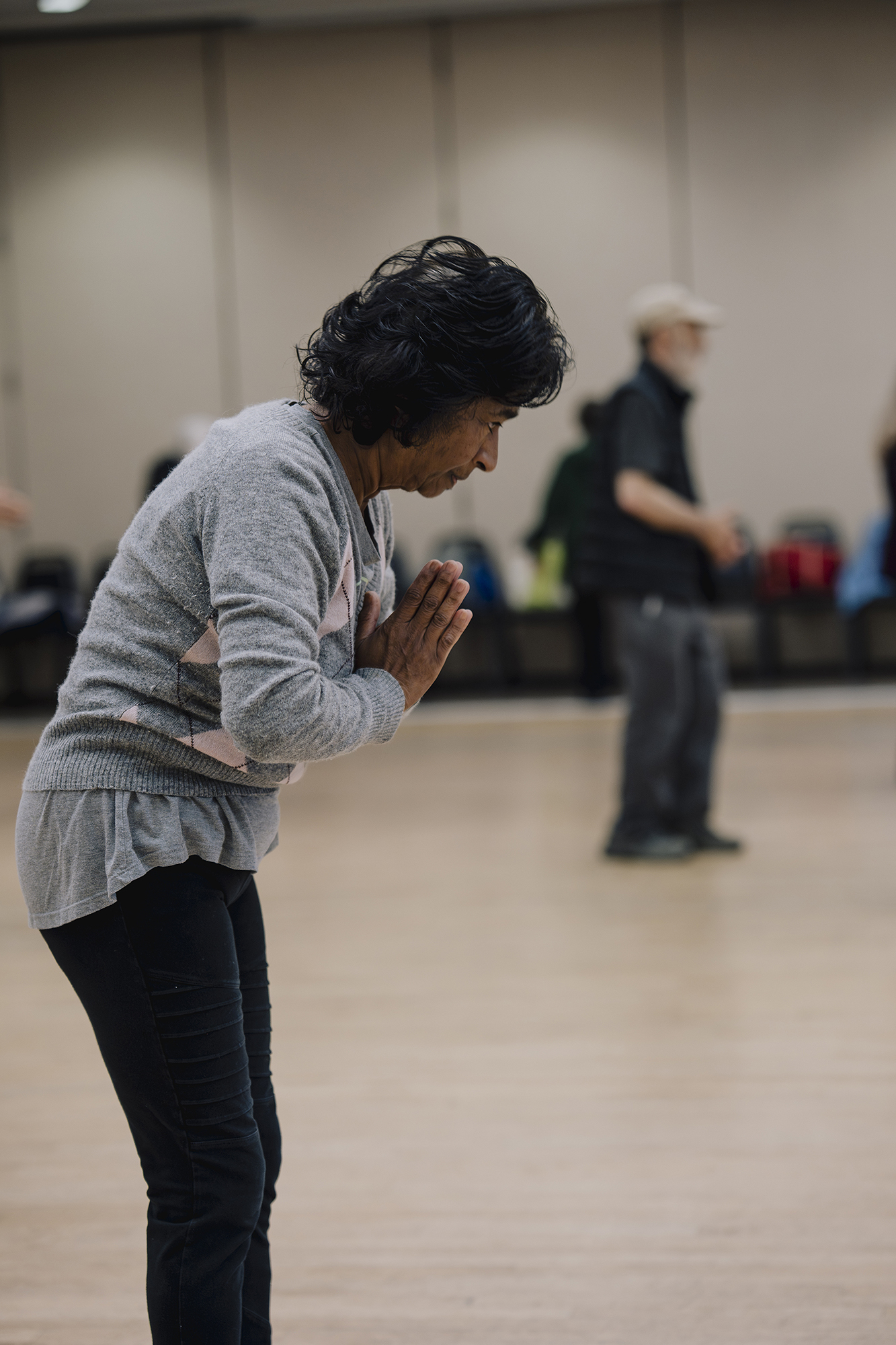 A person stands indoors with hands pressed together and head bowed slightly, while another person stands blurred in the background of a large room.