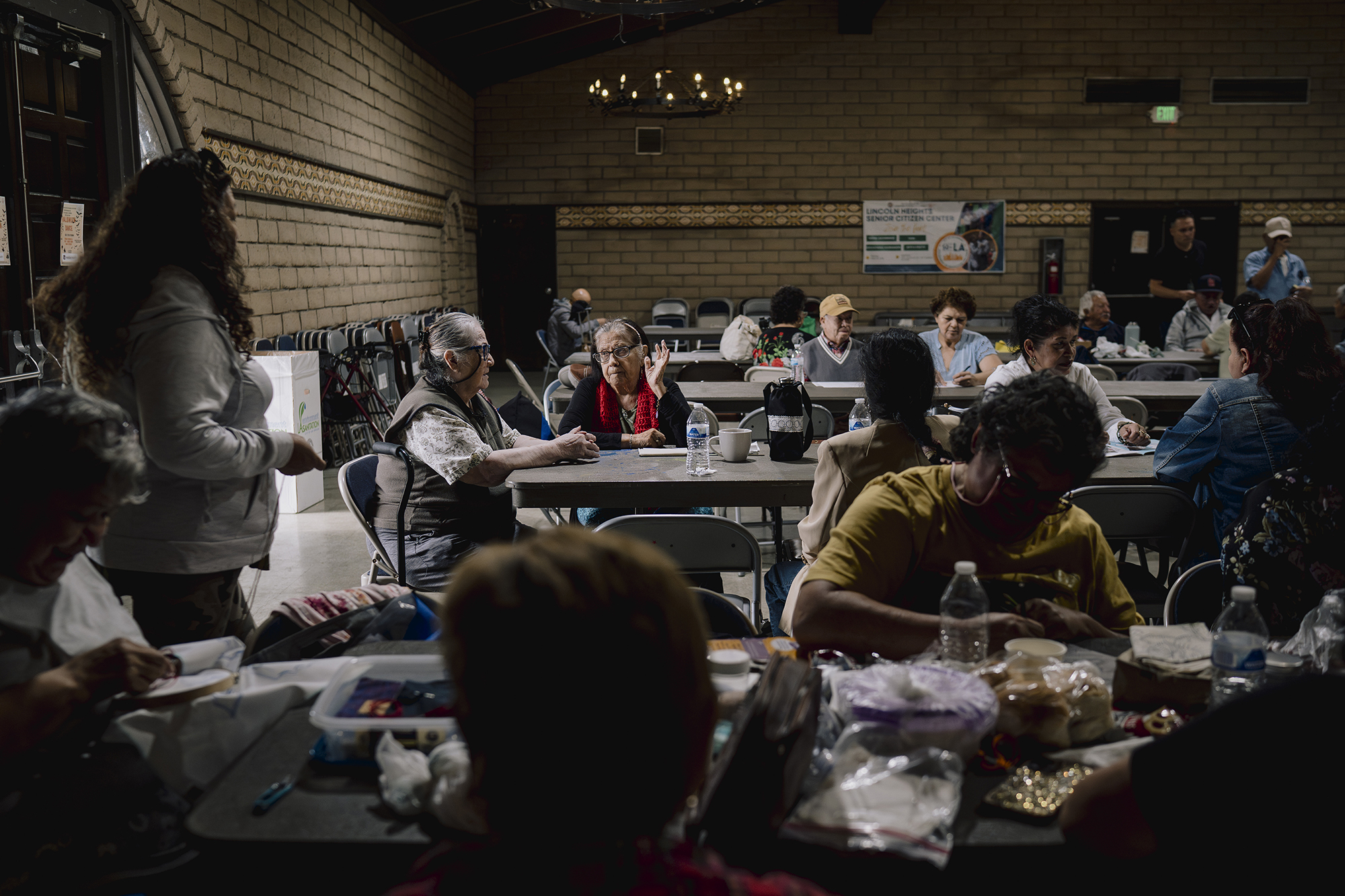 People sit and talk around tables inside a senior center hall, with craft supplies, water bottles, and snacks spread out under warm overhead lighting.