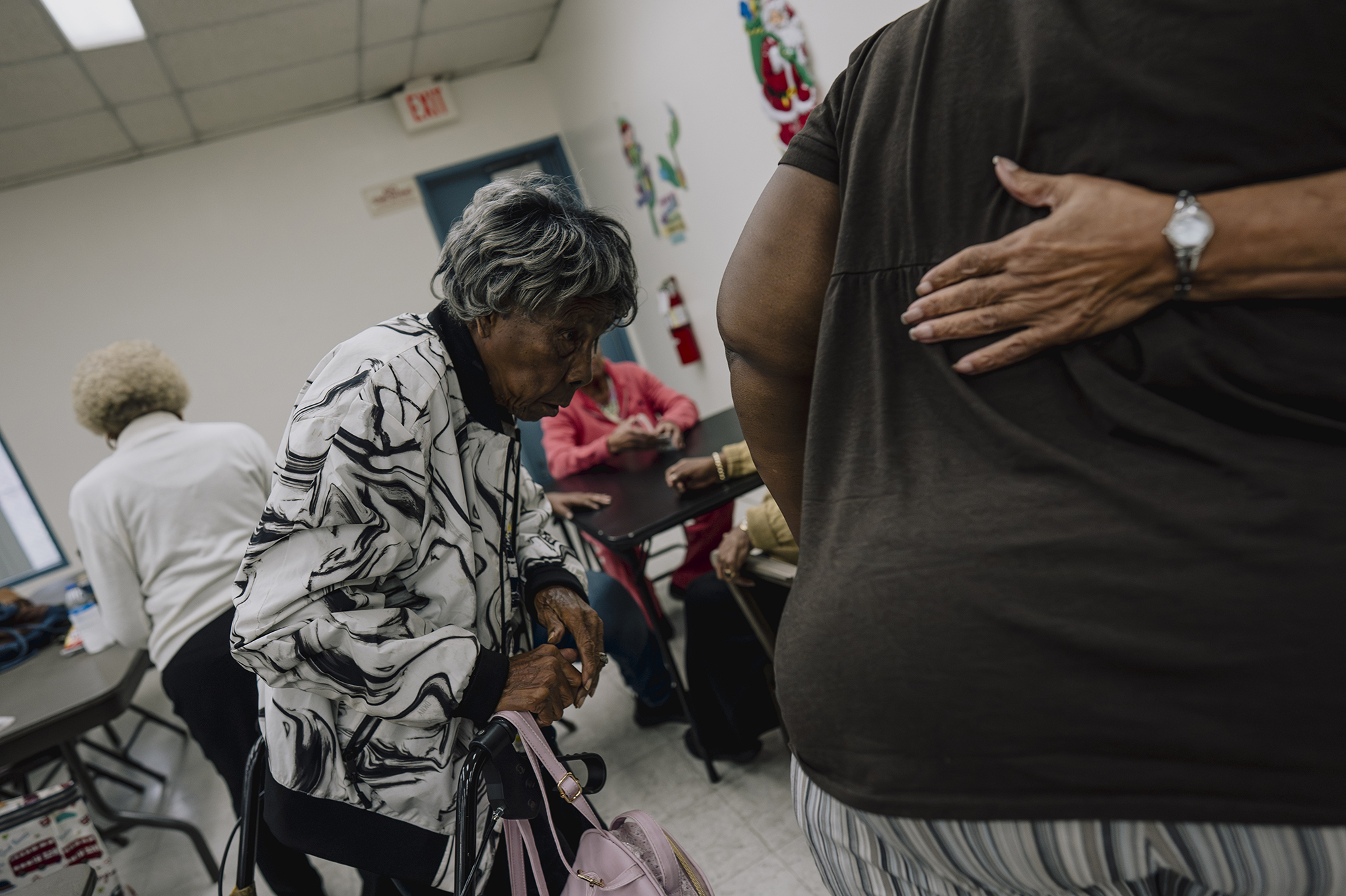 An older adult with gray hair steadies themself on a walker while holding a pink handbag inside a room where several people sit at tables, as another person stands nearby with a hand resting on their back.