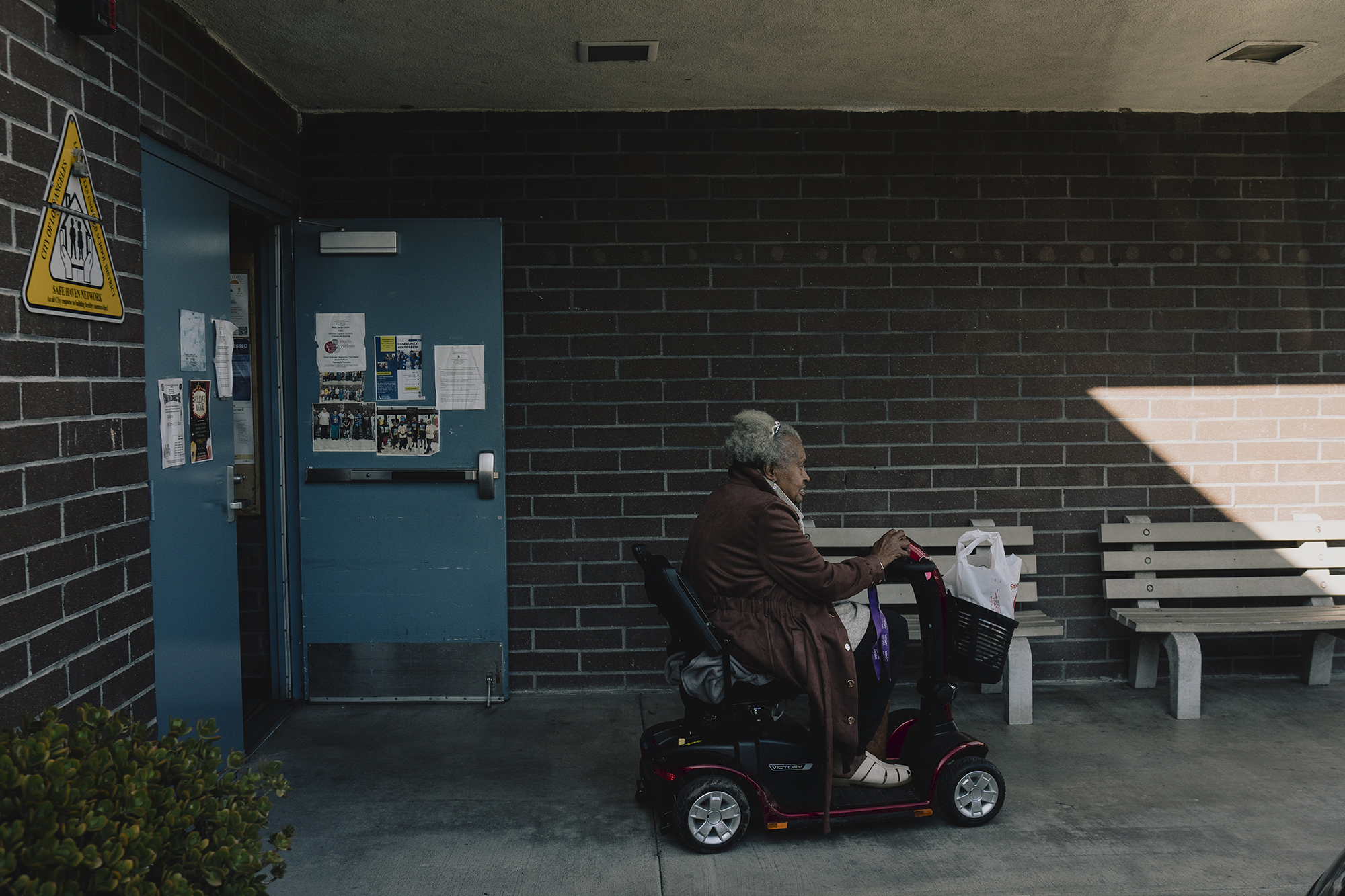 An older adult rides a mobility scooter past a brick wall outside a building entrance, carrying a white plastic bag in the front basket as sunlight falls across part of the wall and bench nearby.