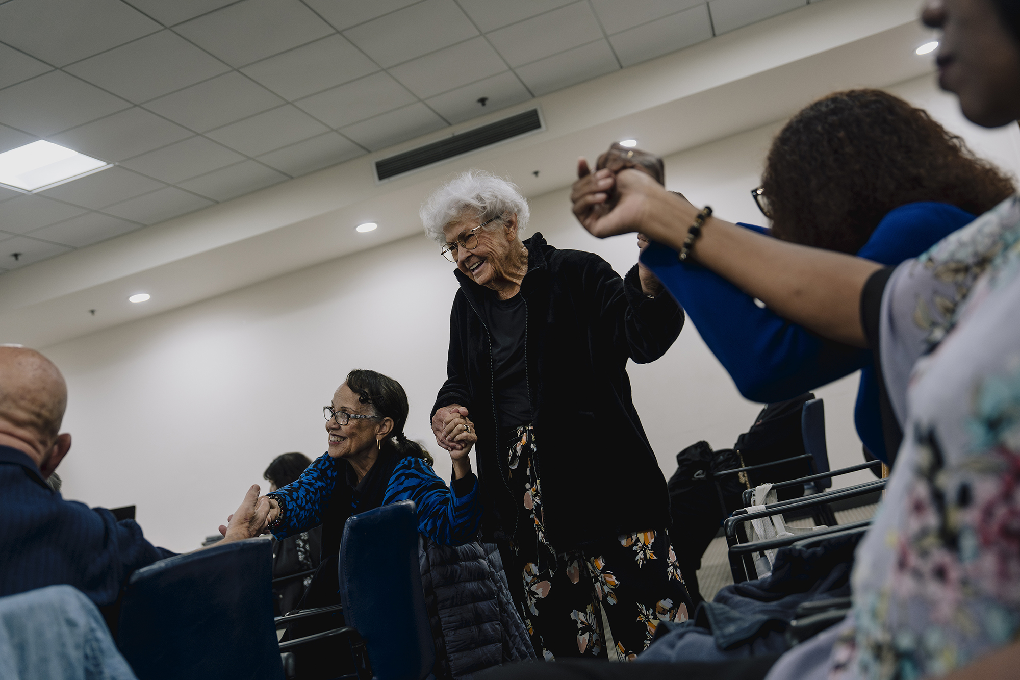 An older adult with white hair smiles while standing and holding hands with others in a circle inside a bright room, as several seated participants reach toward one another in a group activity.