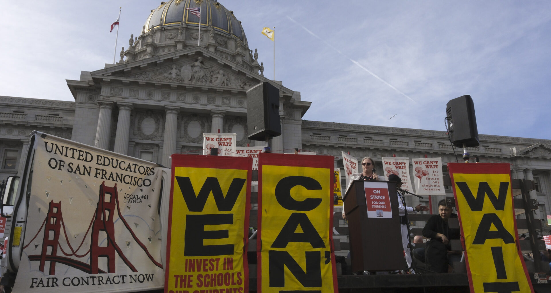 A teacher addresses a rally outside San Francisco City Hall with banners reading "We Can't Wait" and "Invest in the schools our students deserve.
