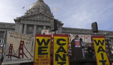 A teacher addresses a rally outside San Francisco City Hall with banners reading "We Can't Wait" and "Invest in the schools our students deserve.