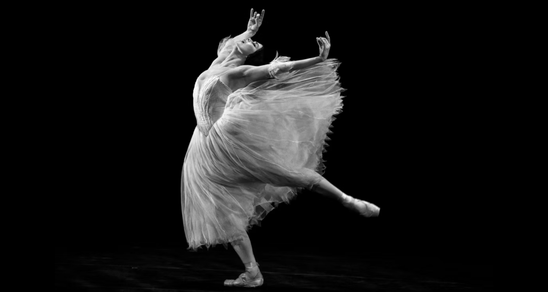 A black and white photo of Natalia Osipova, who dances with abandon onstage. She leans forward with one leg lifted in a low arabesque, her supporting leg slightly bent, as she arches and lifts her arms up and back. Her eyes closed, she opens her mouth with emotion. She wears a Romantic tutu with wispy arm puffs; the skirt of the tutu flies out behind her.