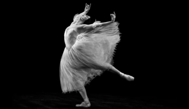 A black and white photo of Natalia Osipova, who dances with abandon onstage. She leans forward with one leg lifted in a low arabesque, her supporting leg slightly bent, as she arches and lifts her arms up and back. Her eyes closed, she opens her mouth with emotion. She wears a Romantic tutu with wispy arm puffs; the skirt of the tutu flies out behind her.