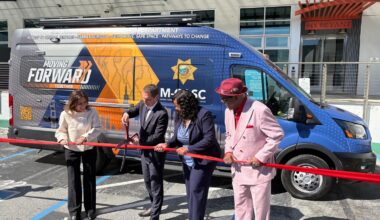 Four people cut a red ribbon in front of a van labeled "Moving Forward" during a ribbon-cutting event in a parking lot.
