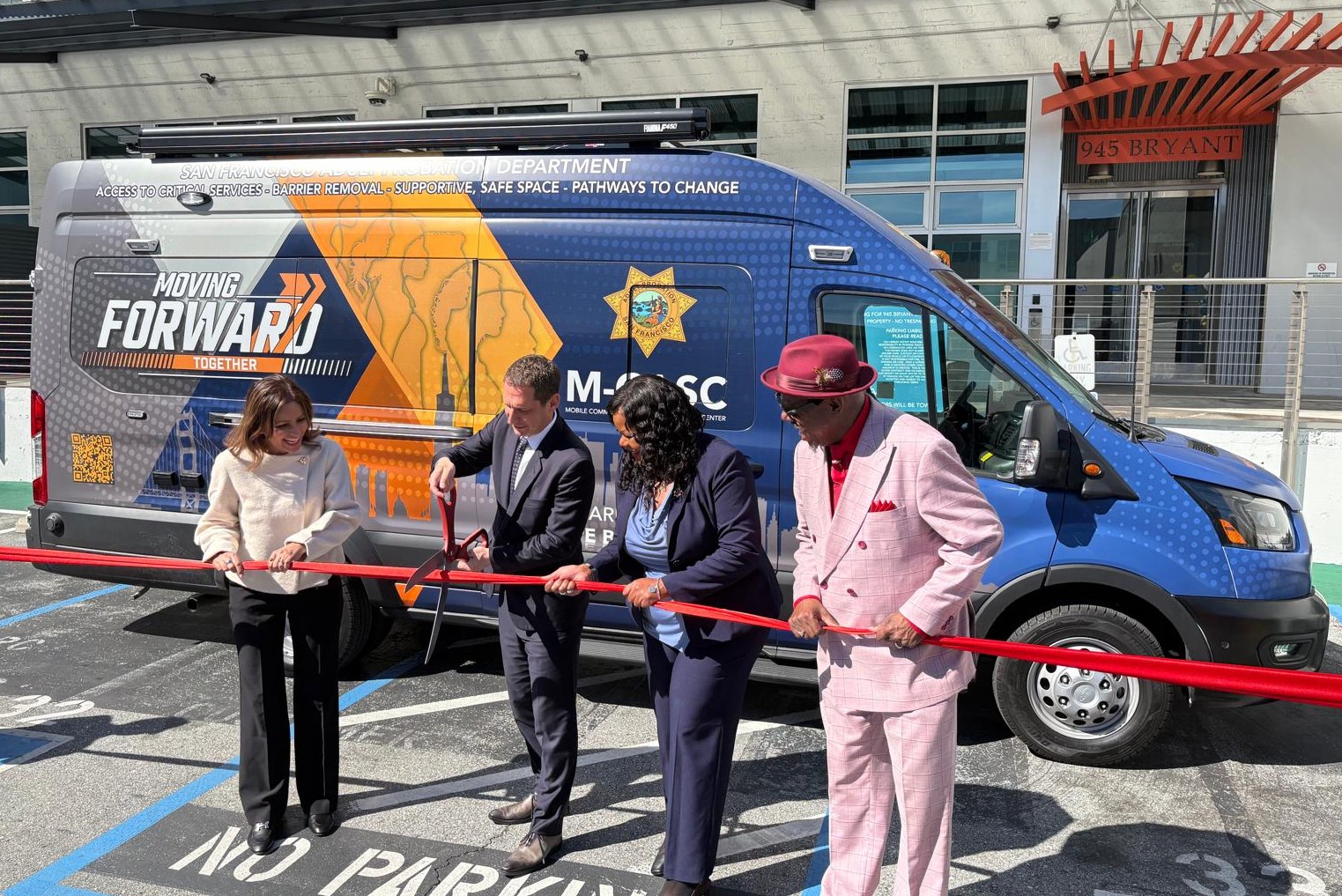 Four people cut a red ribbon in front of a van labeled "Moving Forward" during a ribbon-cutting event in a parking lot.