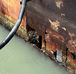 Severely rusted and corroded metal wall above waterline with large holes and exposed interior, next to a black hose.