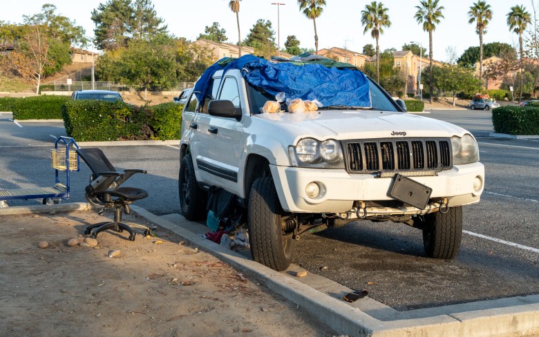 A car partially covered in a tarp parked next to an office chair. Loaves of bread sits on the hood and miscellaneous trash can be seen below the passenger side doors.