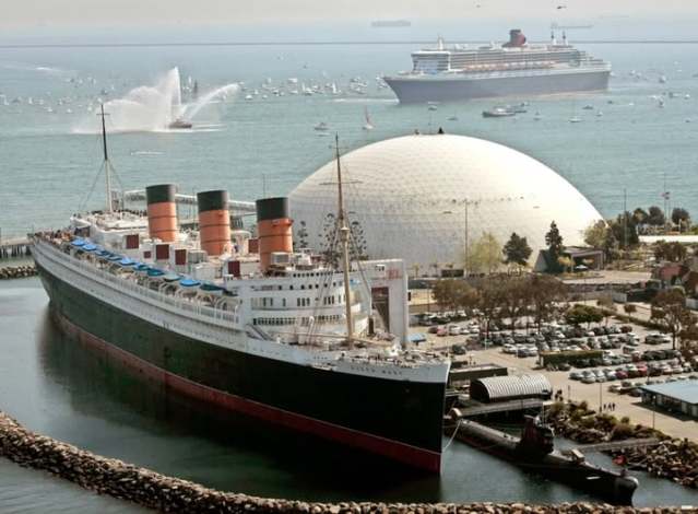 Aerial view of a historic ocean liner docked near a dome-shaped building, with a modern cruise ship in the background and a busy harbour filled with boats.