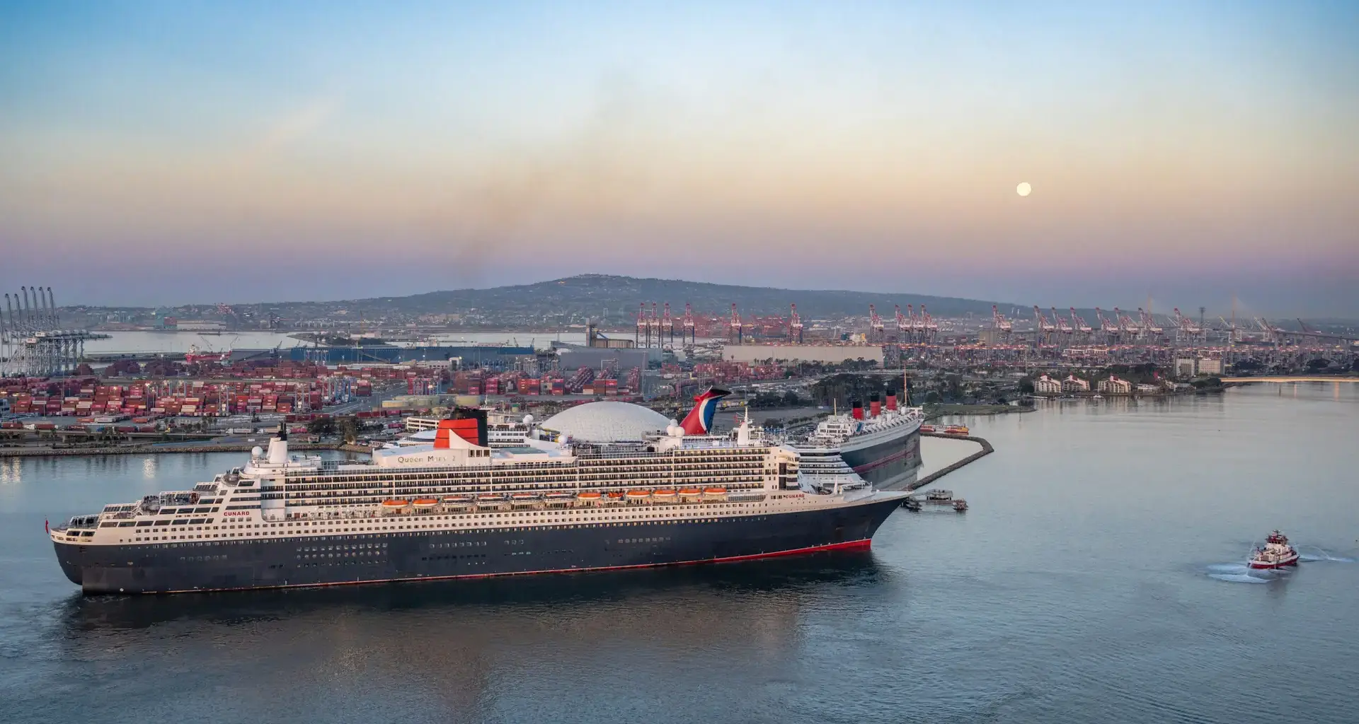 A large cruise ship is docked in a port with shipping containers, cranes, and buildings in the background. The sky is clear with a faint moon visible, and a small tugboat is in the water nearby.