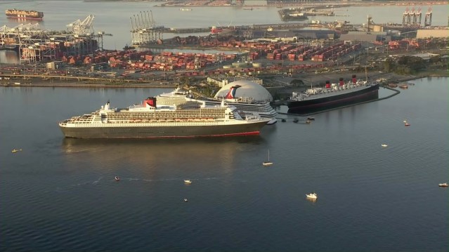 Aerial view of a busy port with two large cruise ships docked in the foreground, surrounded by small boats. In the background, cargo containers and cranes are visible, highlighting the bustling maritime activity.