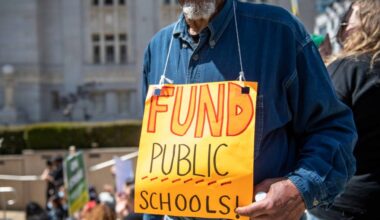 An older Black man stands outside at a rally, with a sign hanging from his neck that says: 'Fund Public Schools.'