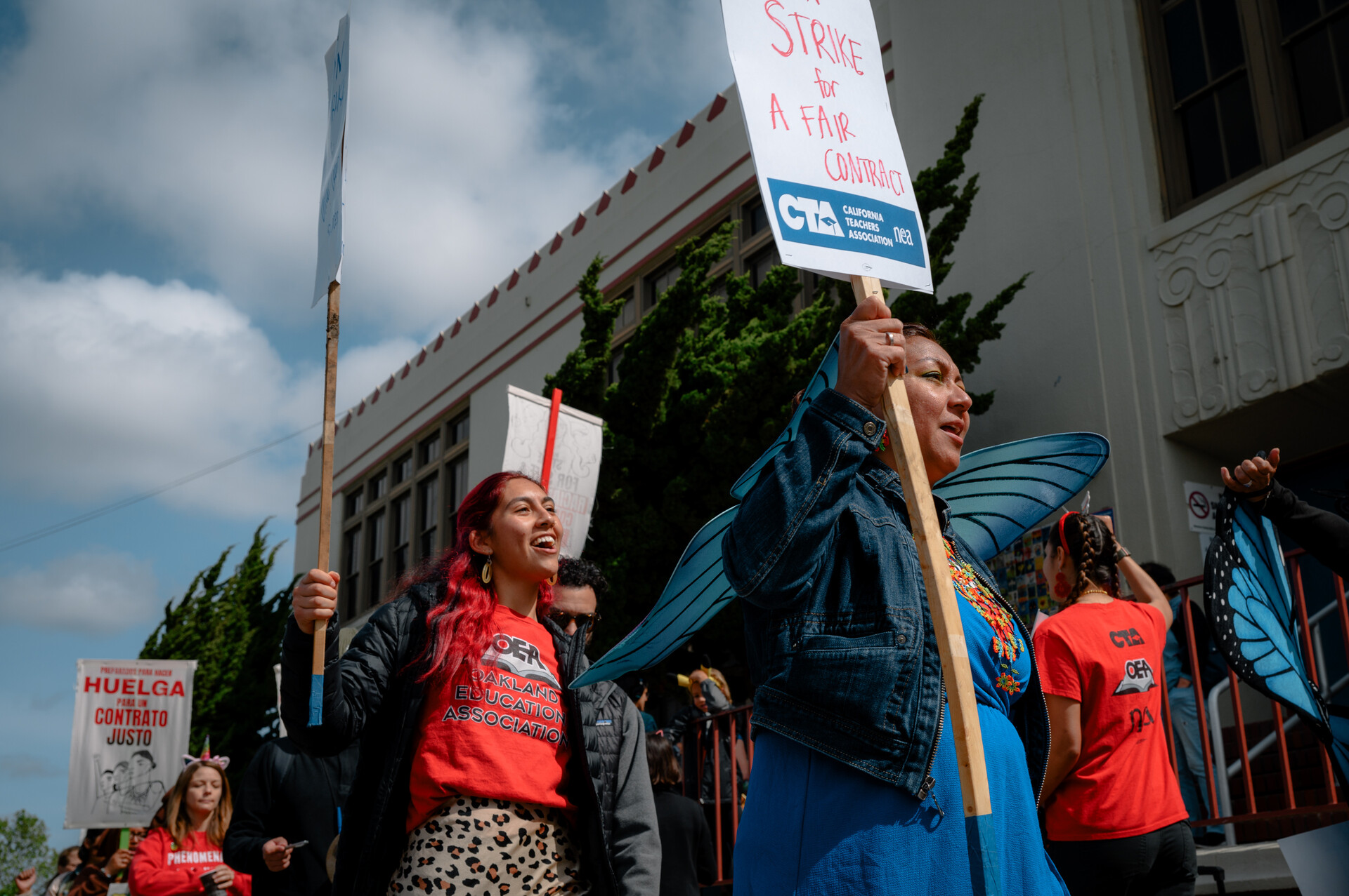 Teachers march in front of a school, holding protest signs.