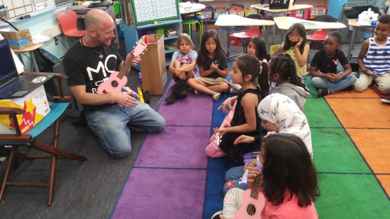 A man teaches children to play pink ukuleles in a classroom setting.