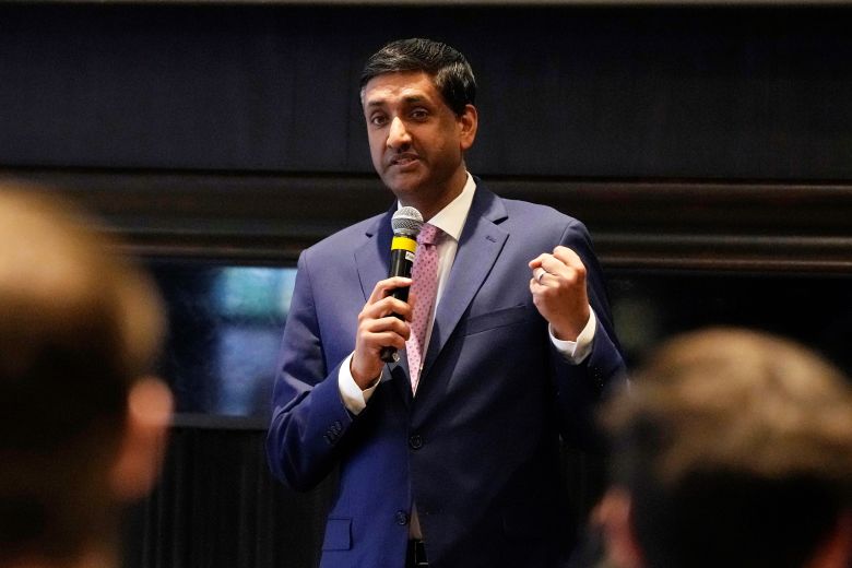 Man in a dark blue suit holding a microphone while speaking at an indoor event.