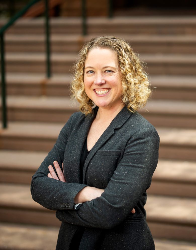Woman with curly blonde hair smiling confidently, arms crossed, wearing a dark blazer. She stands in front of blurred stair steps, conveying a professional tone.