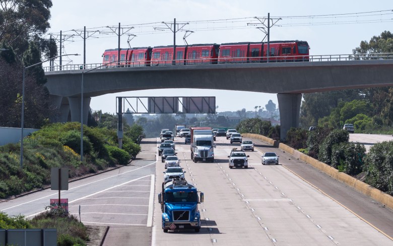 A trolley car is seen on an overpass passing over a multi-lane freeway in the bright daylight.