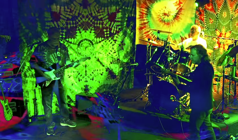 Three men perform rock music against a psychedelic background