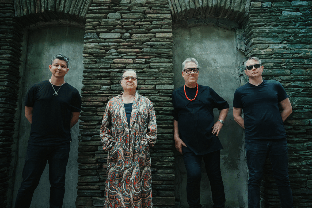 Four male band members pose in front of a stone wall