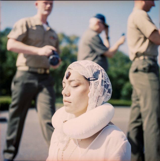 A person with closed eyes and a lace headscarf sits wearing a travel pillow and a stone on their forehead, reminiscent of quiet moments found in museums; two uniformed men stand behind, one holding a camera.