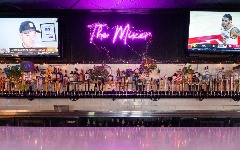A neon purple lit bar with a wall of taps, TVs with sports broadcasts, and a glowing sign reading "The Mixer."