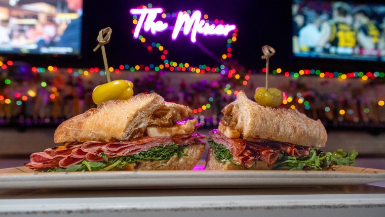 A salami sandwich sits on a bar. A neon sign reading "The Mixer" is seen in the background.