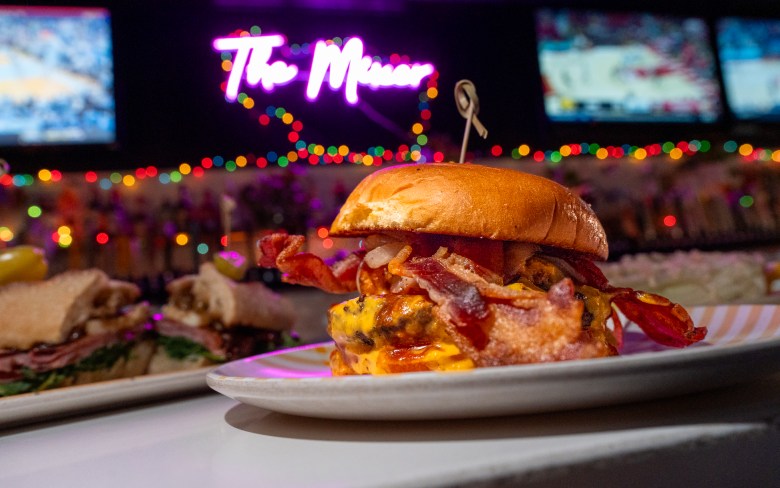 A burger sits on a bar. A neon sign reading "The Mixer" is seen in the background.