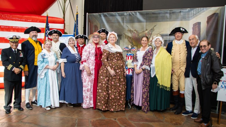 A group photo of individuals in modern and 18th-century-style clothing with an American flag backdrop.