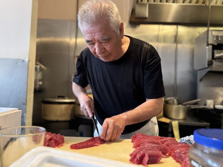 An elderly man in a black shirt slices raw fish on a cutting board in a commercial kitchen.