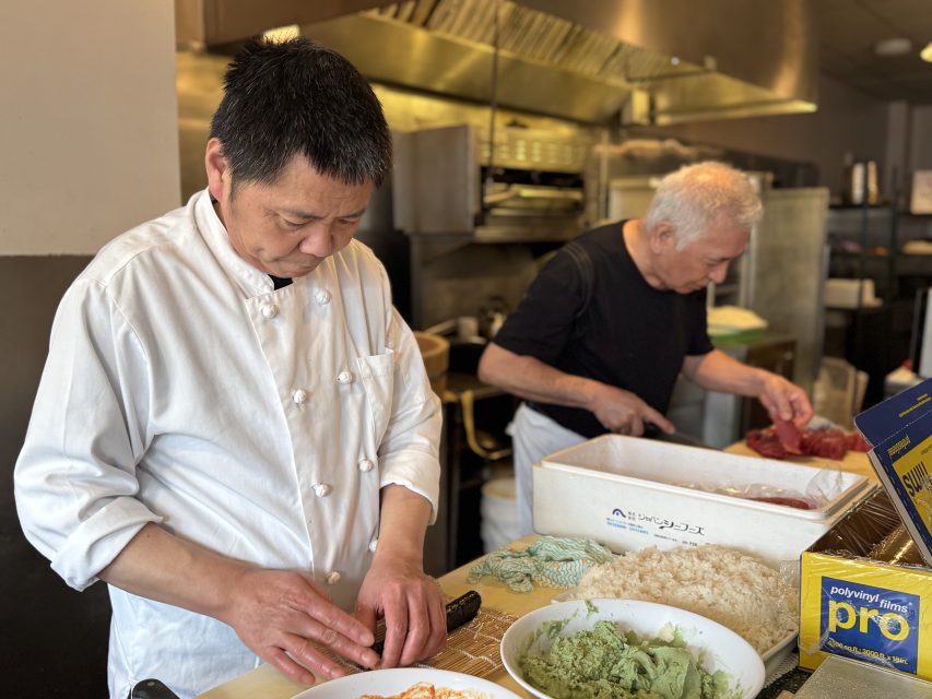Two chefs prepare food in a restaurant kitchen; one is slicing ingredients while the other arranges food on a counter near wasabi and a box of plastic wrap.