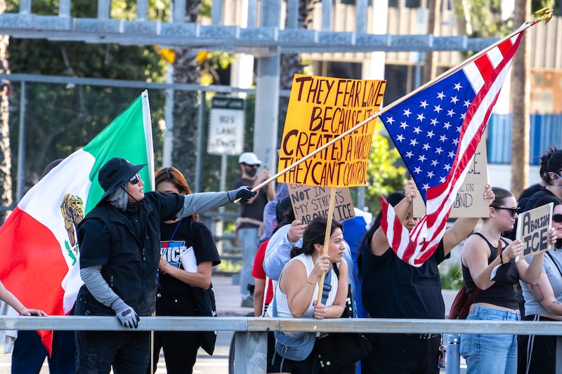 A protester in a black hat and jacket waves an American flag in front of a Mexican flag held by another protester. A woman next to the protester holds a sign reading "They fear love because it creates a world they can't control."
