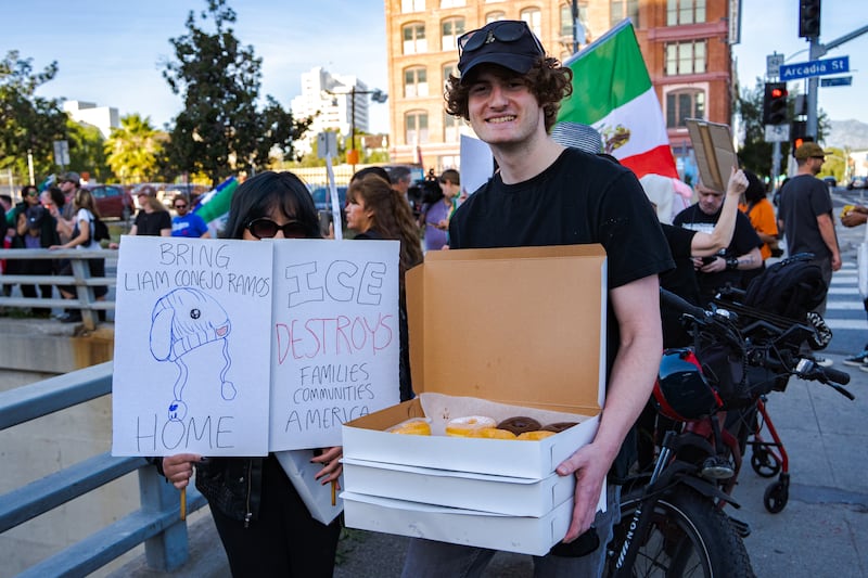 A man holds three boxes of donuts, smiling to the camera, standing next to a woman in sunglasses holding two signs reading "Bring Liam Conejo Ramos home" and "ICE destroys families communities America."