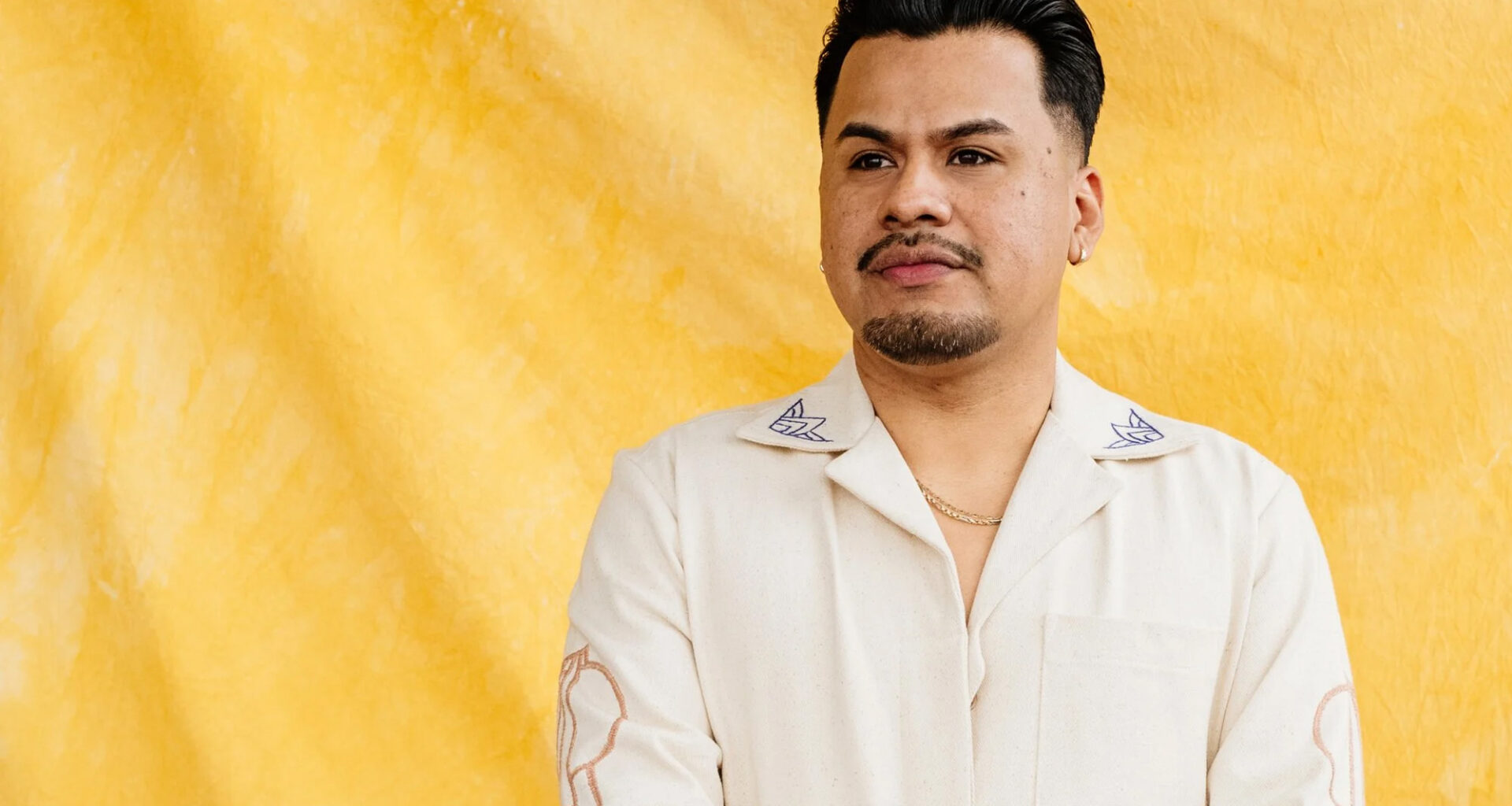 A man in a white suit with patterned decor stands against a yellow backdrop.