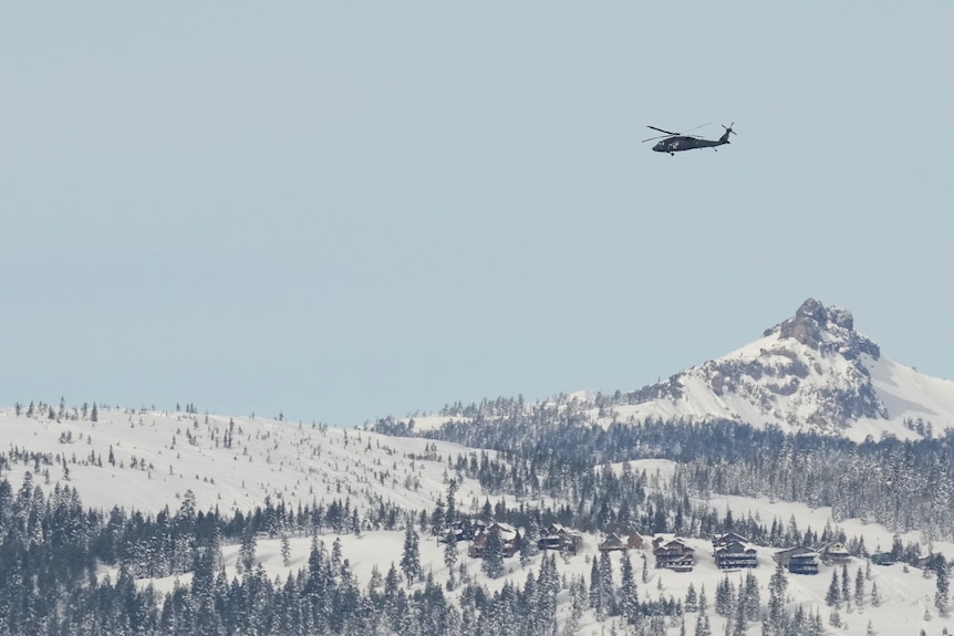 A snowy forested landscape his a black helicopter flying in the distance 
