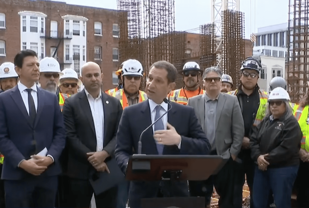 Mayor Daniel Lurie speaking at a podium with District 5 Supervisor Bilal Mahmood and other individuals in hard hats behind him at a construction site.