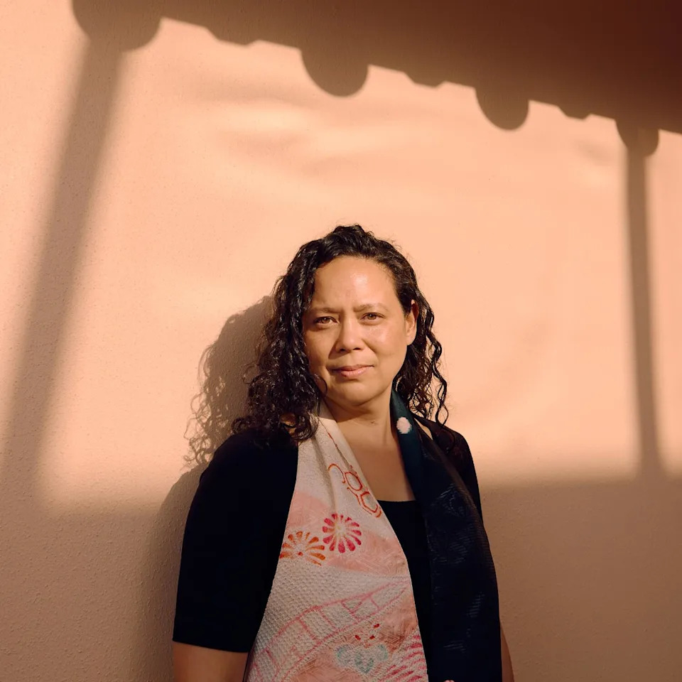 A woman with long black hair, black T-shirt and a shawl poses against a wall framed by shadows.