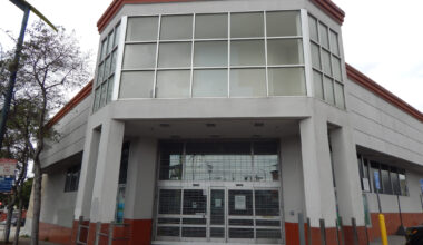 A two-story, gray and red commercial building with large windows and a closed metal security gate over the entrance.