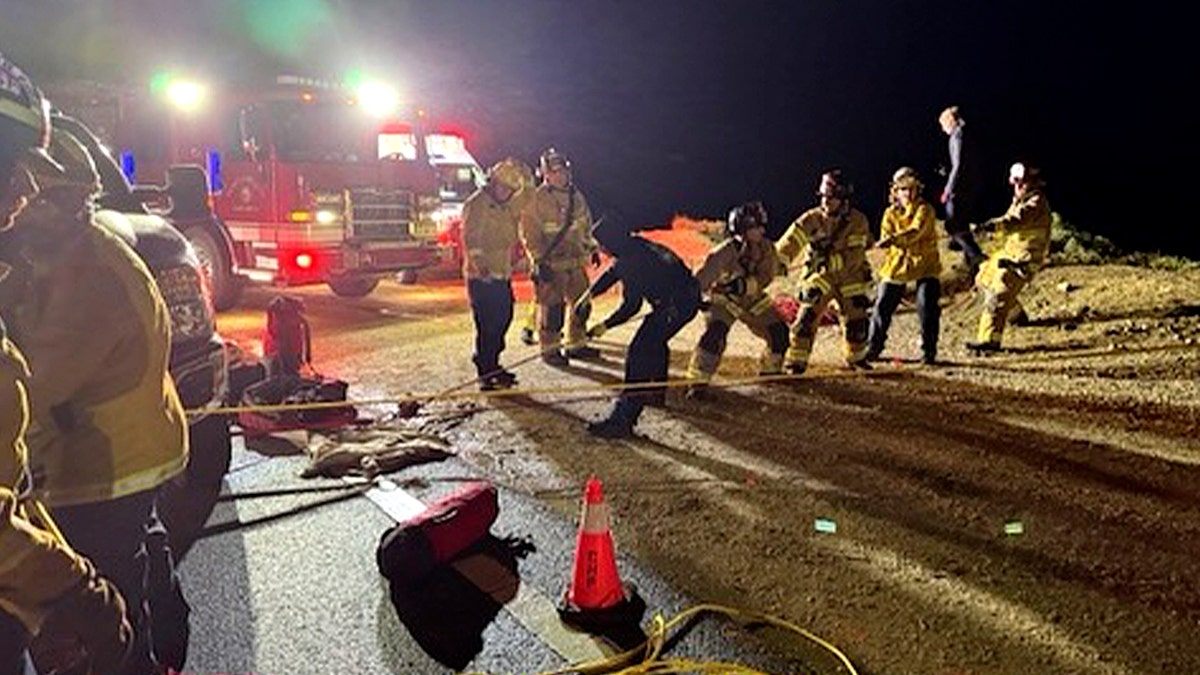 First responders work near a cliffside roadway at night with a fire engine and rescue equipment visible.