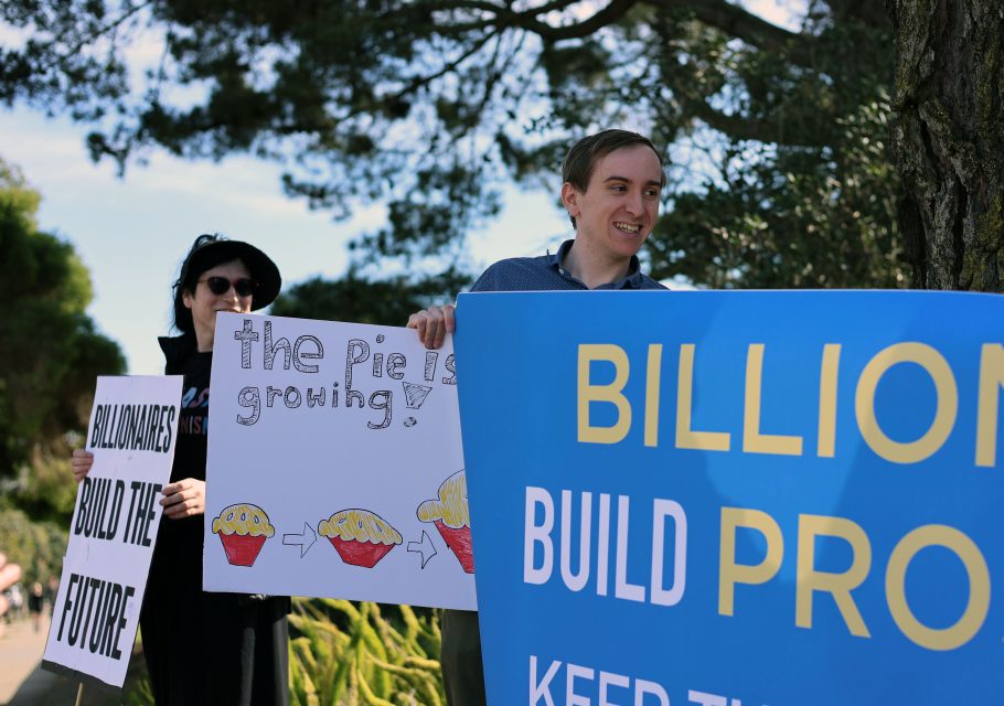 Two people join a billionaire march, holding protest signs about billionaires and economic growth—one sign features pies, while another boldly says "BILLION..." as they stand outdoors near trees.