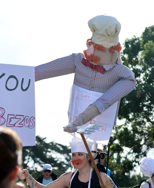 A person in a chef's hat carries a large chef puppet wearing a sign at an outdoor billionaire march, with protest signs visible in the crowd.