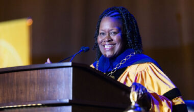 Cal State LA President stands at a podium in academic regalia, smiling while speaking during a ceremony.