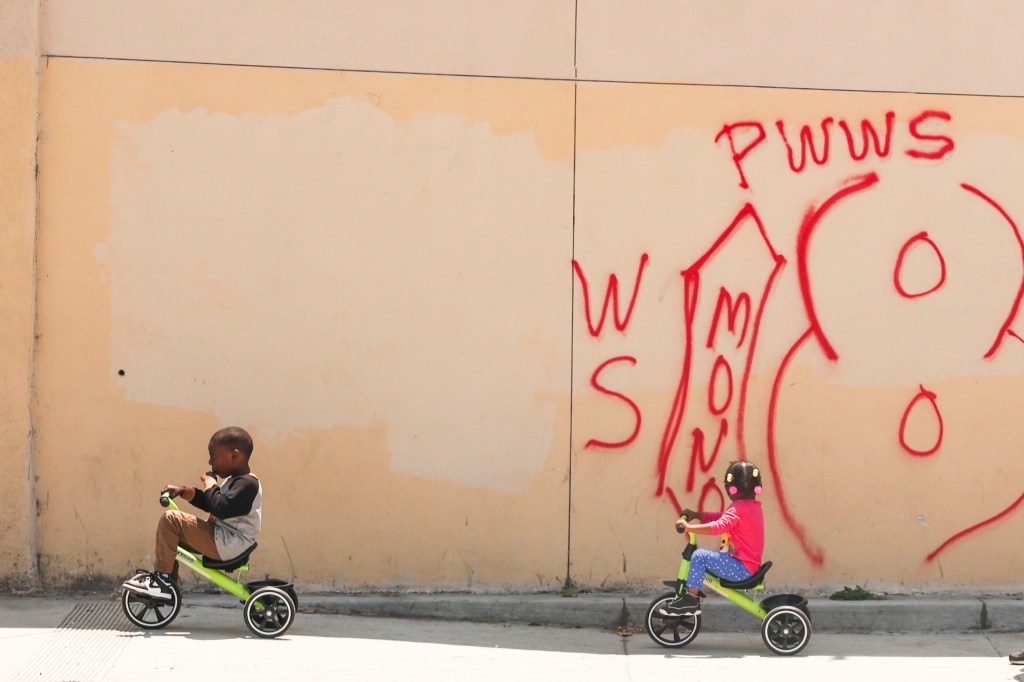 Two children riding bicycles in front of a wall with red gang graffiti in South Los Angeles.