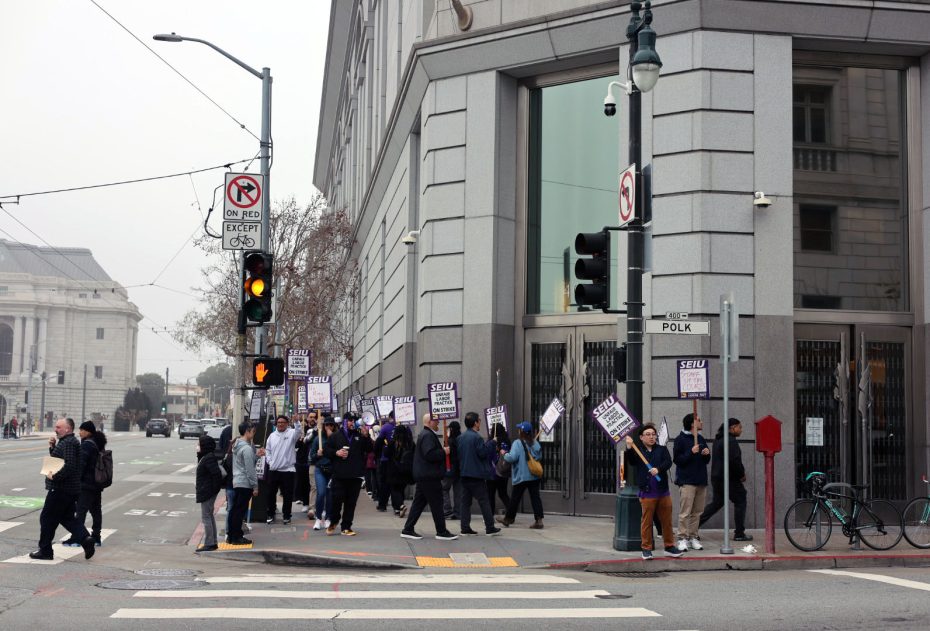 A group of people holding signs march on a city sidewalk at an intersection near a large building on a cloudy day.