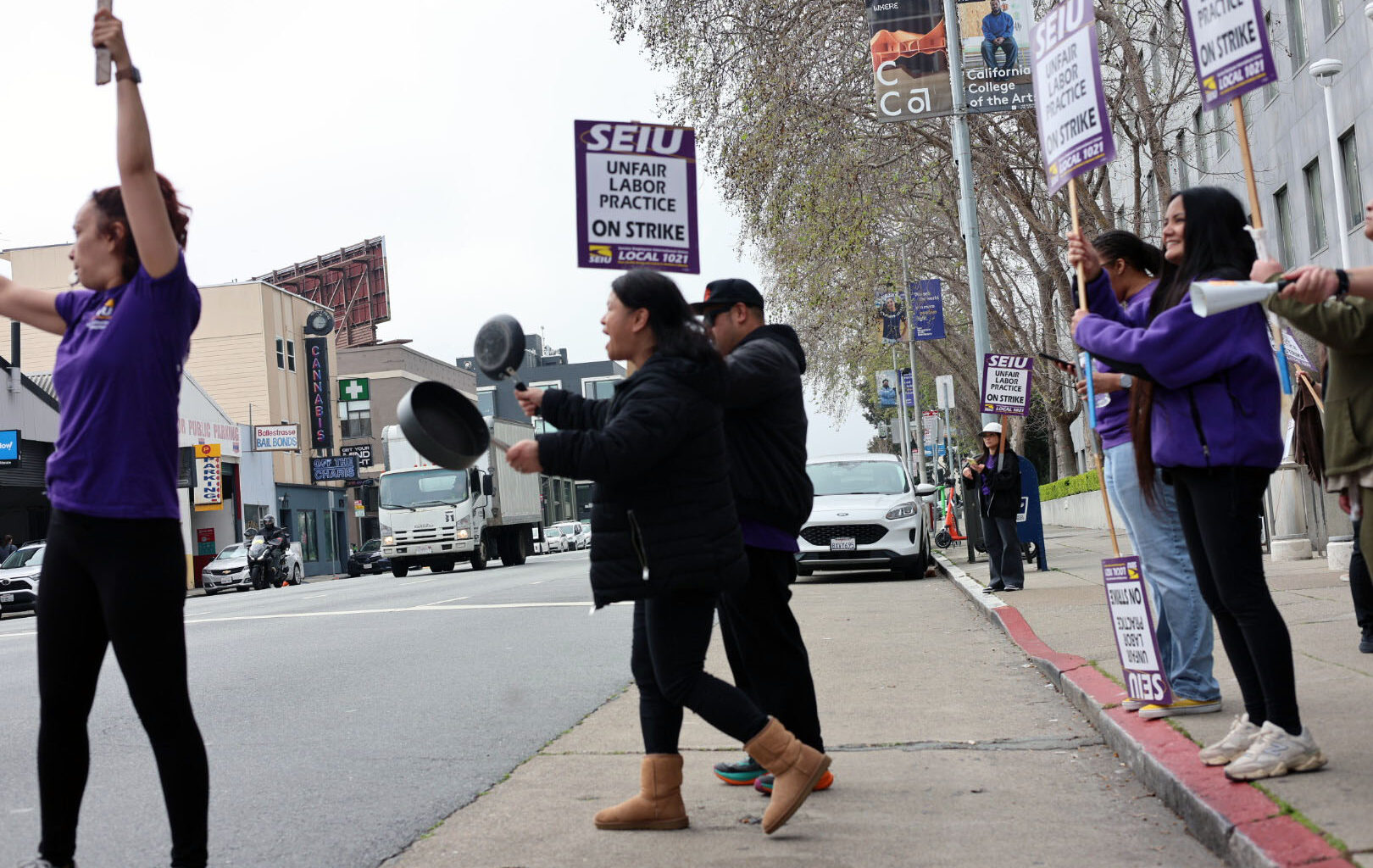 A group of people on a city sidewalk hold SEIU strike signs and bang pots, protesting unfair labor practices.